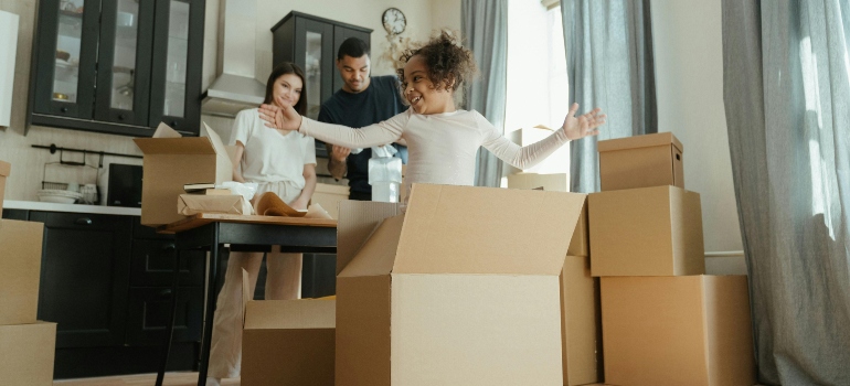 A cheerful child playing with moving boxes while her parents smile in the background.