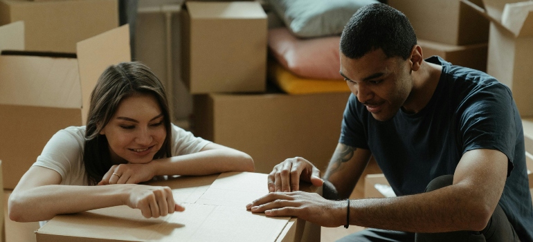 A couple sitting and smiling while packing a cardboard box, representing teamwork when moving your family from a house to an apartment.