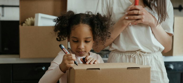 A young girl carefully labeling a cardboard box while her parent supervises, illustrating tips for organized family moves.