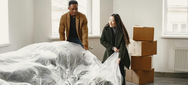 A man and a woman draping protective plastic over a sofa inside a sparsely furnished room.