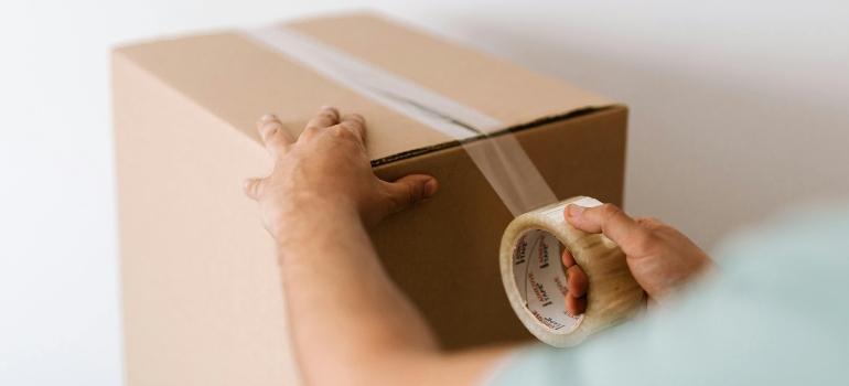 A close-up of a hand applying tape on a sealed cardboard box against a plain white wall.