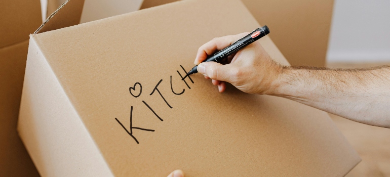 Close-up of a person writing 'Kitchen' on a cardboard box to organize moving items.