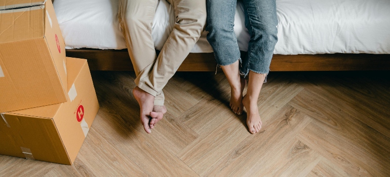 Couple sitting on the edge of a bed, their feet dangling over the floor, surrounded by neatly packed moving boxes.