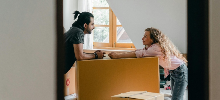 Two people smiling and discussing plans while leaning on a cardboard box in their new home.