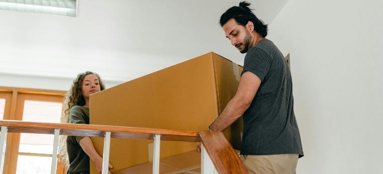 Two people carefully lifting a large cardboard box together on a staircase while moving from an apartment to a house.