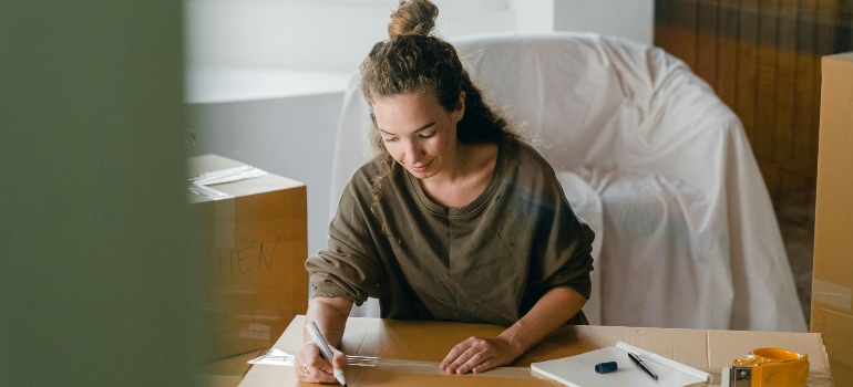 A woman seated at a table writing labels on moving boxes for her home relocation.