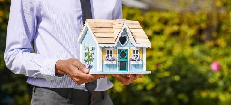 A person holding a miniature blue house model, representing the dream of moving from an apartment to a house. 