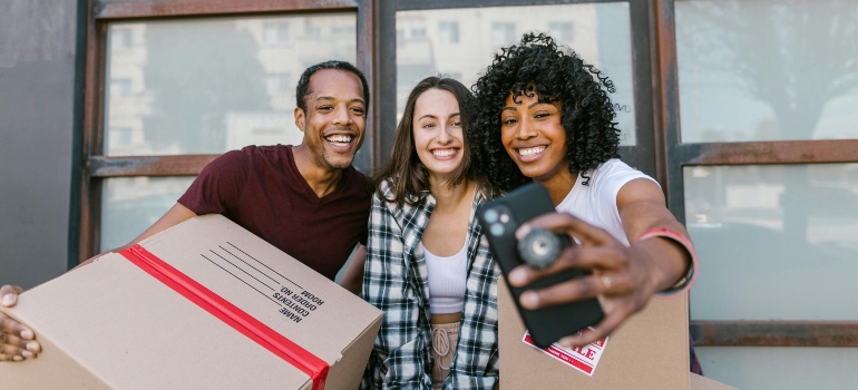 Three friends hold boxes and capture a selfie after a successful moving day.