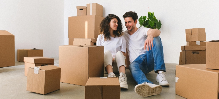 A smiling young couple relaxing on a floor, surrounded by stacked cardboard boxes in a bright room.