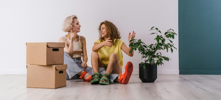 Two smiling friends sitting on the floor, surrounded by boxes, enjoying a relaxed moment during their move.