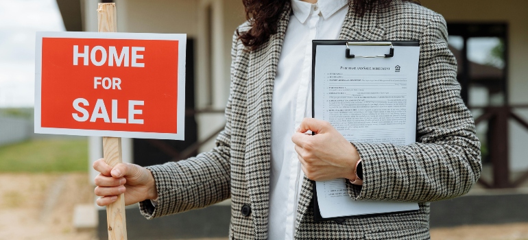 "A real estate agent holding a clipboard and a 'Home for Sale' sign in front of a property.