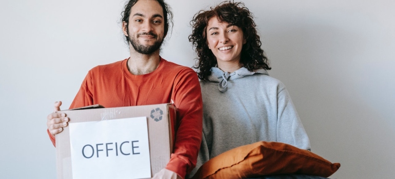 a man holding a box labeled 'office' and a woman holding a pillow before relocating a home and family business simultaneously